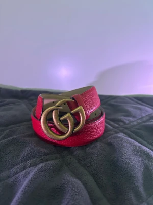 Rött skärp från Gucci med GG-spänne - Snyggt rött skärp från Gucci i skinn med det ikoniska dubbla GG-spännet i antikguld. Bältet har en klassisk design med justerbara hål och en lyxig känsla. Perfekt accessoar för att lyfta vilken outfit som helst. Längden är 100cm. Helt ny