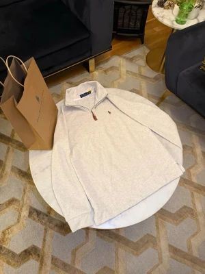 Ljusgrå stickad tröja Polo Ralph Lauren - Ljusgrå stickad tröja från Polo Ralph Lauren med halv dragkedja och klassisk broderad logga på bröstet. Tröjan har hög krage och ribbade muddar. Perfekt för lager-på-lager och snygg till jeans eller chinos.