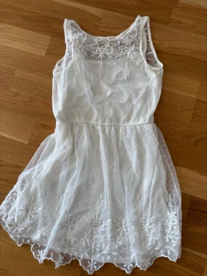 Vit spetsklänning med broderi - Superfin vit kortklänning med broderade blommor och spetsdetaljer. Klänningen är ärmlös och har en lätt, luftig känsla med markerad midja. Perfekt för sommarens alla tillfällen och ger en romantisk vibe.