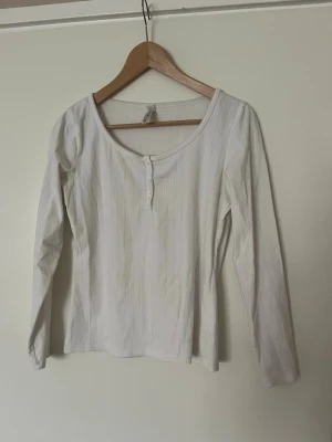 Vit långärmad topp med knappar - En enkel och stilren vit långärmad topp med rund halsringning och små knappar framtill. Toppen är tillverkad i ett mjukt material och har en lätt ribbad struktur och fint mönster som ger en snygg touch. 