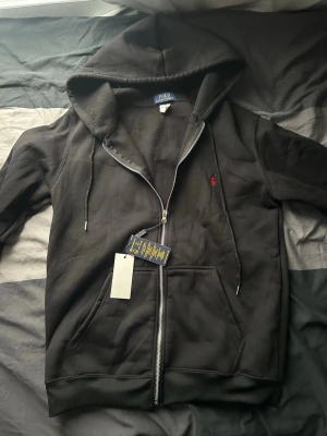 Svart hoodie från Polo Ralph Lauren - Svart hoodie med dragkedja från Polo Ralph Lauren. Klassisk design med huva, dragsnören och känguruficka framtill. Liten röd logga broderad på bröstet. Mjuk bomullsblandning som är skön att ha på sig. Perfekt för en chill och stilren look.