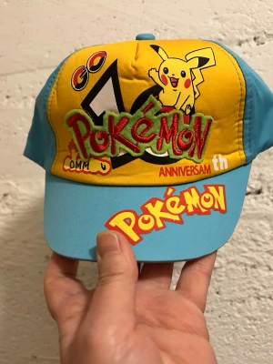Pokémon keps med Pikachu och logga - Färgglad keps med gul front och blå sidor samt skärm. Framsidan har Pikachu och Pokémon-logga i brodyr och tryck, samt text om jubileum. Justerbar rem baktill. Perfekt för Pokémon-fans som vill sticka ut. Stlk 2-4 år, helt ny 