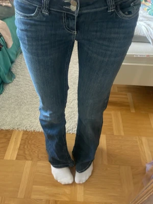 Blå bootcut jeans med kontrastsömmar - Säljer ett par blå bootcut jeans med vita kontrastsömmar på bakfickorna och dubbla knappar i midjan. Jeansen har klassisk femficksdesign och är tillverkade i stretchigt denimtyg för skön passform. Perfekta till sneakers eller boots.