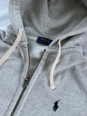 Polo Ralph Lauren Zip Sweater - Snygg grå hoodie från Polo Ralph Lauren med dragkedja, huva med dragsko och två fickor framtill. Klassisk logga broderad på bröstet. Perfekt för en avslappnad och stilren look.