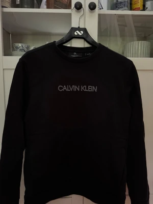 Svart Calvin Klein sweatshirt - Svart sweatshirt från Calvin Klein med broderad logga på bröstet. Tröjan har rund halsringning och långa ärmar. Enkel och stilren design, perfekt för en avslappnad look.