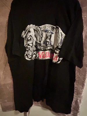 Svart Valhalla t-shirt  - Cool svart t-shirt med stort Valhalla-tryck framtill. Motivet har vikingainspirerade detaljer i grått, vitt och rött. T-shirten har en klassisk passform och korta ärmar, perfekt för dig som gillar mytologi och streetstyle. Stlk kvar L, XL och XXL , 120kr/st , helt ny , samma frakt 