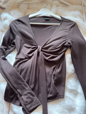Brun knyttopp från Gina Tricot - Snygg brun topp från Gina Tricot med lång ärm och knyt framtill. Toppen har en djup v-ringning och är tillverkad i mjukt bomullsmaterial. Perfekt för att skapa en trendig och avslappnad look.