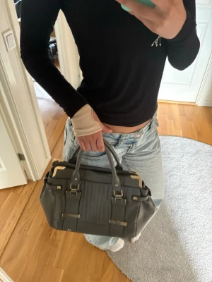 Mörkgrå handväska med gulddetaljer - Snygg mörkgrå handväska med två handtag och coola guldfärgade metalldetaljer. Så snygg väska med gulddetaljer. På vissa ställen är guldet inte lika synligt pga användningen💗💗