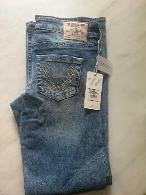 True Religion ljusblå jeans med strass - Ljusblå jeans från True Religion med låg midja och bootcut-modell. Bakfickan har en snygg strassdetalj i form av märkets ikoniska båge. Klassisk denim med patch och etikett bak. Perfekta för dig som gillar statement-jeans. Dessa är samarbetet med Tilda Törnqvist. Supersnygga! Små i storleken, så passar garanterat en 27a också. 