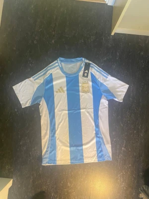 Argentina Adidas fotbollströja blå/vit - Säljer en officiell Argentina fotbollströja från Adidas med klassiska blå och vita vertikala ränder. Tröjan har korta ärmar, rund hals och guldiga detaljer samt AFA-emblem på bröstet. Perfekt för dig som älskar fotboll och vill visa ditt stöd för landslaget.