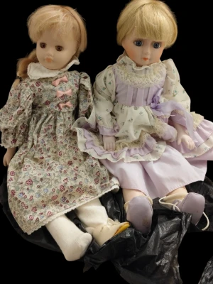 Två porslinsdockor i vintagekläder - Säljer två porslinsdockor med blommiga och pastellfärgade klänningar, spetsdetaljer och rosetter. Dockorna har blont hår och vita strumpor, samt skor i lila och beige. Perfekta som dekoration eller samlarobjekt.