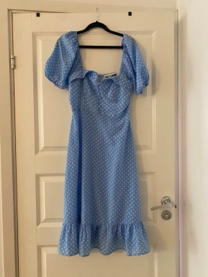 Blå prickig klänning från Boohoo - Supersöt blå klänning från Boohoo med vita prickar, puffärmar och volang nertill. Klänningen har knappar framtill, söt knytning vid bysten och dragkedja i ryggen. Perfekt för sommarens alla tillfällen!