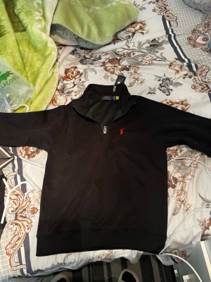 Svart half-zip tröja Polo Ralph Lauren - Snygg svart half-zip tröja från Polo Ralph Lauren med klassisk röd logga broderad på bröstet. Tröjan har hög krage och ribbade muddar vid ärmslut och nederkant. Perfekt för en stilren och avslappnad look.