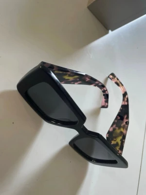 Svarta fyrkantiga solglasögon Prada  - Snygga solglasögon från Prada med svart, fyrkantig båge och trendiga skalmar i camouflage-mönster med inslag av rosa, grön och beige. Perfekt accessoar för att lyfta din stil med en cool och modern touch. Säljes utan låda och tecken på användning finns. 