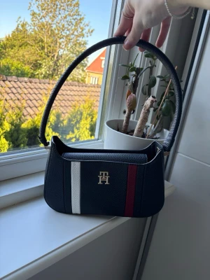 Tommy Hilfiger mörkt blå axelväska - Snygg svart axelväska från Tommy Hilfiger i präglat läder. Väskan har ett stilrent TH-emblem i guld framtill samt dekorativa ränder i vitt och rött. Perfekt storlek för det viktigaste och en klassisk form med kort axelrem. Aldrig använd!