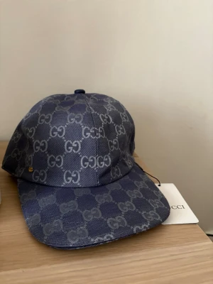 Blå GG-mönstrad keps  - Snygg blå keps från Gucci med klassiskt GG-monogram över hela ytan. Kepsen har böjd skärm och justerbar rem baktill i guldtonad metall. Tillverkad i canvasmaterial med vit insida och märkestaggar kvar.