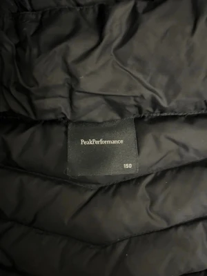 Svart dunjacka Peak Performance - Snygg svart dunjacka från Peak Performance med huva och dragkedja framtill. Jackan har quiltad design och logga på bröstet. Perfekt för kalla vinterdagar och håller dig varm med stil.