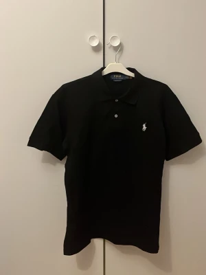 Svart piké från Polo Ralph Lauren - Klassisk svart pikétröja från Polo Ralph Lauren med vit broderad logga på bröstet. Modellen är custom slim fit och har korta ärmar samt knappar vid halsen. Perfekt för en clean och stilren look. Storlek M men passar även S