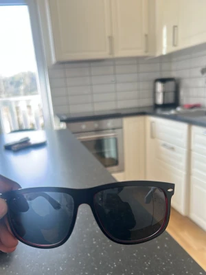 Svarta röda solglasögon med röd detalj - Snygga svarta solglasögon med klassisk wayfarer-form och en tunn röd kant längs insidan av bågarna. Perfekta för att ge din stil en cool touch. Lätta och bekväma att bära hela dagen.