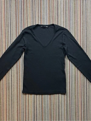 Svart ribbad långärmad topp - Svart långärmad topp med v-ringning och ribbad struktur. Perfekt att styla med jeans eller kjol för en clean look. Enkel och tidlös design som passar till många outfits.
