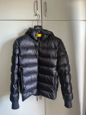 Svart pufferjacka från Parajumpers - Snygg svart pufferjacka från Parajumpers med huva och dragkedja framtill. Jackan har en gul detalj vid halsen och ribbade muddar vid ärmslut och midja. Perfekt för kalla vinterdagar och riktigt skön att bära. Köpt från johnells för 6500-: använd 5 gånger så i nyskick!