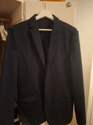 Mörkblå kavaj från Lindbergh White - Stilren mörkblå kavaj från Lindbergh White i slim fit-modell. Klassisk design med smala slag, tre fickor framtill och enkelknäppning. Perfekt för dig som gillar en modern och snygg look. Materialet känns mjukt och följsamt.