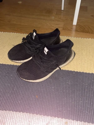 Svarta Adidas Ultra Boost sneakers - Säljer ett par svarta Adidas Ultra Boost sneakers med stickad ovandel och vita Boost-sulor. Skorna har klassisk låg profil, svarta snören och diskret Adidas-logga på plösen. Perfekta för dig som gillar stilrena och bekväma sneakers.
