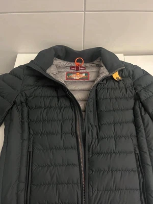 Svart dunjacka Parajumpers herr - Snygg svart dunjacka från Parajumpers med quiltad design och grått foder. Jackan har hög krage, dragkedja framtill och två sidofickor med dragkedja. Klassisk gul detalj vid halsen och orange hängare. Perfekt för kyliga dagar.