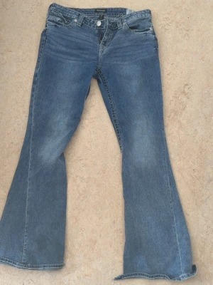 Blå utsvängda jeans med rå kant - Säljer ett par blå jeans från True religion med utsvängda ben och råa kanter nertill. Jeansen har klassisk flappocket och är tillverkade i denim. Lite slitna på botten!