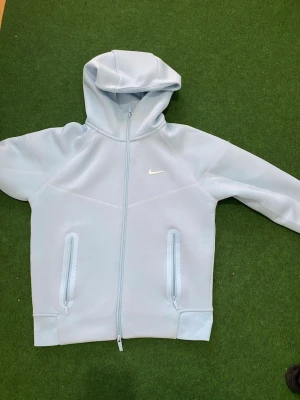 Ljusblå Nike hoodie med dragkedja - Snygg ljusblå hoodie från Nike med hel dragkedja framtill, två fickor med dragkedja och klassisk huva. Diskret Nike-logga på bröstet. Perfekt för en sportig och avslappnad stil.