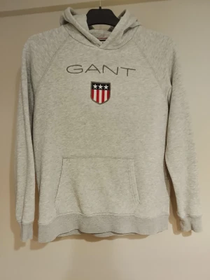 Grå hoodie från GANT med logga NY! - Snygg ljusgrå hoodie från GANT med broderad logga och sköld på bröstet. Klassisk känguruficka framtill och huva. Mjuk bomullsblandning som är perfekt för en avslappnad stil. helt ny, mycket trendig & det finns inte många av dom, först till kvarn!
