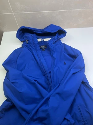 Blå vindjacka från Polo Ralph Lauren - Snygg blå vindjacka från Polo Ralph Lauren med huva och dragkedja framtill. Jackan har en klassisk passform och är tillverkad i ett lätt, vindtätt material. Ikoniska Polo-loggan broderad på bröstet. Perfekt för blåsiga dagar. Är lx i barn så motsvarar S
