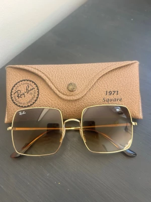 Ray-Ban Square RB1971 914751 54 - Ray-Ban 1971 Square solglasögon med stora fyrkantiga glas och tunn guldfärgad metallbåge. Glasen är bruna med en snygg toning. Kommer med original Ray-Ban-fodral i brunt läderlook. Perfekt accessoar för en trendig look.                                             • BÅGFORM: Rektangulär • BÅGFÄRG: Blank Arista Guld • BÅGMATERIAL: Metall • SKALMFARG: Arista Guld