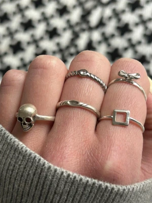 Set med fem silver ringar - Säljer ett set med fem silvriga ringar i olika designer. En ring har en dödskalle, en har tvinnad design med liten sten, en har en rosett, en är fyrkantig och en har ett ovalt mönster. Perfekt för dig som gillar unika detaljer och mixade stilar.