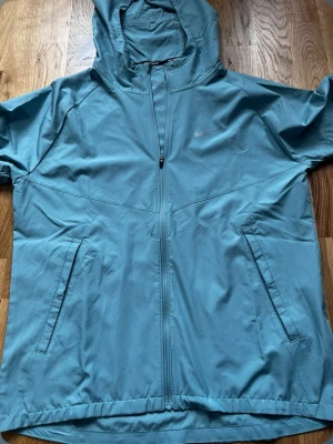 Turkos nike windbreaker/jacka - ALDRIG ANVÄND, Snygg  turkos vindjacka från Nike med hel dragkedja, två snedställda fickor och huva. Jackan har en sportig look och är tillverkad i ett lätt material som passar perfekt för blåsiga dagar. Diskret Nike-logga på bröstet. MÖTS HELT UPP I HELAINGBROG 