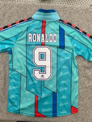 Retro Barcelona fotbollströja Kappa - Snygg retro Barcelona fotbollströja från Kappa med Ronaldo och nummer 9 på ryggen. Tröjan är turkos med röda och blå detaljer, klubbmärke på bröstet och Kappa-loggor längs ärmarna. Tillverkad i lätt och ventilerande syntetmaterial, perfekt för fotboll eller samling.