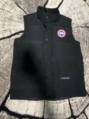 Svart dunväst från Canada Goose - Snygg svart dunväst från Canada Goose med klassisk logotyp på bröstet och hög krage. Västen har tryckknappar framtill och två stora fickor. Perfekt för lager-på-lager och håller dig varm under kyliga dagar.