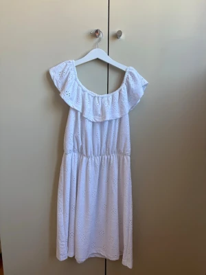 Vit sommarklänning med volang - Söt vit klänning med broderade detaljer och volang vid axlarna. Klänningen har en avslappnad passform och resår i midjan. Perfekt för varma dagar och har en romantisk känsla med sin ärmlösa design.