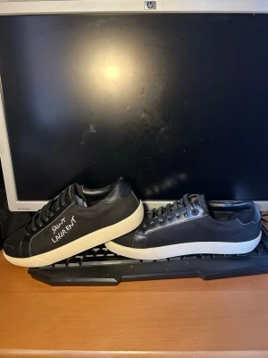 Svarta Saint Laurent sneakers - Svarta sneakers från Saint Laurent med vit logga på sidan. Skorna har lågt skaft, svarta skosnören och vit sula. Tillverkade i en mix av läder och textil för en clean och stilren look, använda endast på en bröllop en gång sedan har dem varit i boxen, först till kvarn