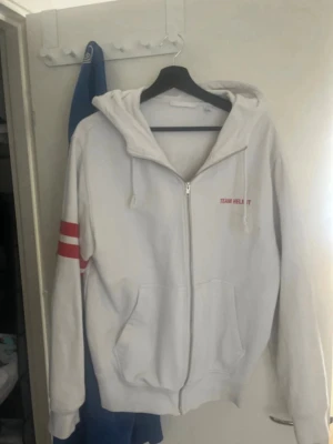 Vit Helmut Lang hoodie med tryck - Vit hoodie med dragkedja från Helmut Lang. Stor röd logga och text på ryggen, samt röda ränder på ena ärmen. Huva med dragsko och fickor framtill. Perfekt för en clean och sportig look.
