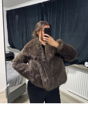 Brun fuskpälsjacka med krage - Säljer en fluffig brun fuskpälsjacka med bred krage och långa ärmar. Jackan har en boxy passform och stängs framtill. Perfekt för dig som vill ha en trendig och varm look under vintern.