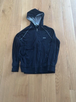 Svart hoodie från BOSS med dragkedja - Snygg svart hoodie från BOSS med hel dragkedja framtill och huva med grått innerfoder. Diskret BOSS-logga på bröstet och vita detaljer längs axlarna. Perfekt för en avslappnad och stilren look.