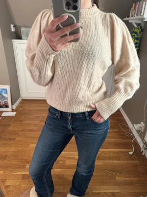 Beige stickad tröja med puffärm - Mysig beige stickad tröja med ribbat mönster och trendiga puffärmar. Tröjan har rund halsringning och långa ärmar med breda muddar. Perfekt för dig som gillar en stilren och modern look.