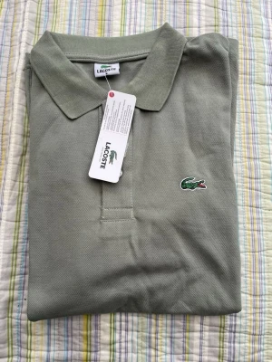 Grön pikétröja från Lacoste - Klassisk ljusgrön pikétröja från Lacoste med krage och knappslå framtill. Tröjan har det ikoniska krokodilmärket broderat på bröstet och är tillverkad i bomull. Perfekt för en stilren och avslappnad look.