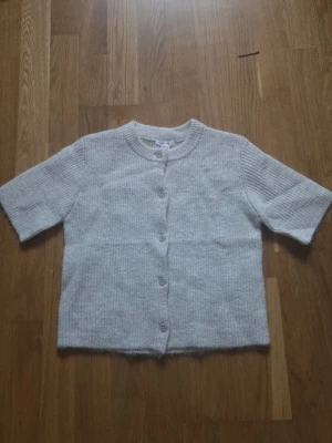 Ljusgrå stickad kofta med kort ärm - Mysig ljusgrå stickad kofta med korta ärmar och rund hals. Koftan har knappar framtill och är gjord i ett mjukt, ribbat material. Perfekt att slänga över en topp för en avslappnad look. Har aldrig använt, ny utan prislapp.