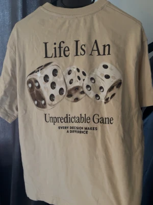 Beige t-shirt med tärningsmotiv - Beige t-shirt från Zara med tryck av tre tärningar och texten 'Life Is An Unpredictable Game' på ryggen. Framtill finns samma text i mindre format. Klassisk rund hals och korta ärmar. Skön bomullskvalitet och avslappnad passform. Använd ett fåtal gånger, då den inte passar mig längre. Passformen är lite oversized, riktigt gött till sommarn.
