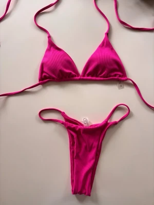 Rosa ribbad bikini med knyt - Snygg rosa bikini i ribbat material med trekants-bh och smala knytband. Helt ny aldrig använd från Shein och köptes för 130kr. Uttagbar vaddering i toppen. 