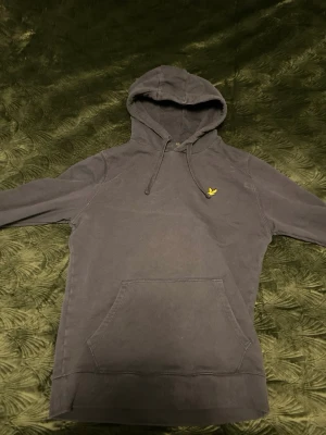 Mörkgrå hoodie från Lyle & Scott - Snygg mörkgrå hoodie från Lyle & Scott med klassisk gul logga på bröstet. Hoodien har huva med dragsko, stor magficka och ribbade muddar vid ärmslut och nederkant. Tillverkad i mjukt bomullsmaterial som är skönt att ha på sig.