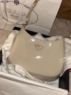 Beige axelremsväska från Prada - Snygg beige axelremsväska från Prada i glansigt material med guldfärgade detaljer och klassisk trekantig logga framtill. Väskan har en stilren, rundad form och justerbar axelrem. Perfekt för dig som vill ha en lyxig och trendig handväska.
