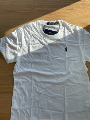 Vit Ralph Lauren T-shirt - 👕 Märke: Polo Ralph Lauren 📏 Storlek: M 🎨 Färg: Vit med svart logga ✨ ☀️ Fräsch och stilren t-shirt från Polo Ralph Lauren – perfekt för sommaren och enkel att matcha med både shorts och jeans. Tillverkad i mjuk bomull med klassisk passform och ikonisk svart logga på bröstet.  ✨ Skick: Helt ny med tags kvar 🧼 Aldrig använd 👌 Enkel, tidlös och riktigt snygg till vardags